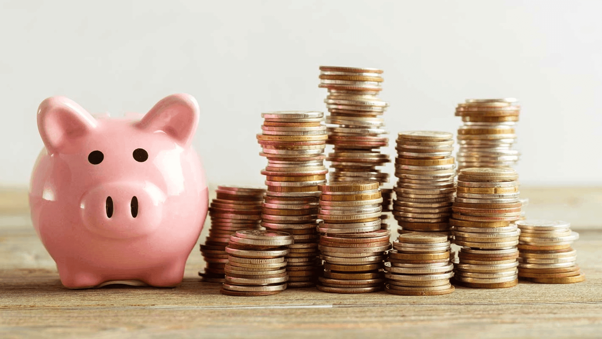 Une tirelire en forme de cochon rose à gauche, avec plusieurs piles de pièces de monnaie empilées à droite, représentant l'épargne et la gestion de l'argent.