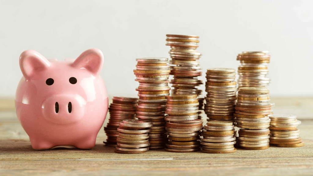 Une tirelire en forme de cochon rose à gauche, avec plusieurs piles de pièces de monnaie empilées à droite, représentant l'épargne et la gestion de l'argent.