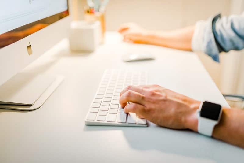 Personne travaillant sur un ordinateur de bureau Mac avec un clavier sans fil, portant une montre connectée au poignet, dans un environnement de bureau lumineux et minimaliste.